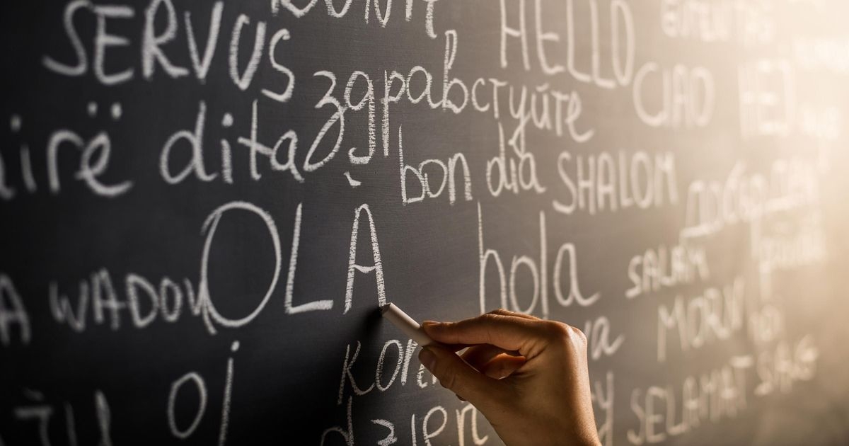 Eine Hand mit einem Stück Kreide an einer Schultafel mit dem Wort Hallo in verschiedenen Sprachen