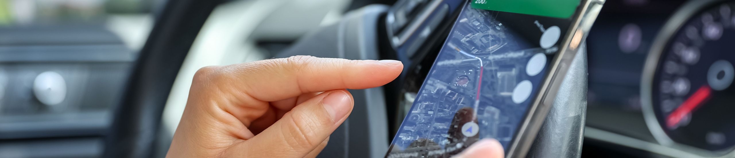 Ein Mensch sitzt in einem Auto. Seine Hand zeigt auf ein Smartphone, das er in der rechten Hand hält. 