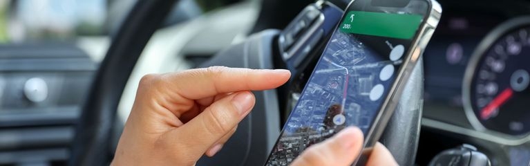 Ein Mensch sitzt in einem Auto. Seine Hand zeigt auf ein Smartphone, das er in der rechten Hand hält. 