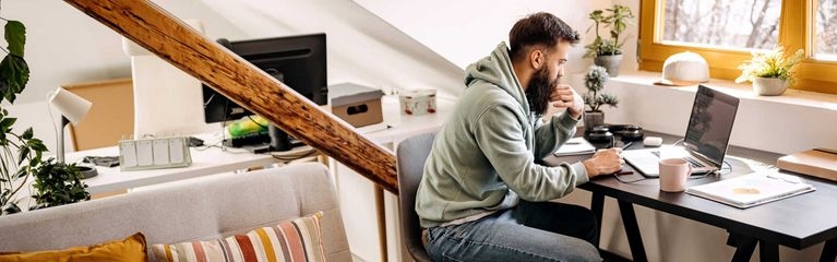 Junger Mann mit Bart sitzt am Schreibtisch und arbeitet am Laptop, neben seinem Stuhl schläft ein Hund.