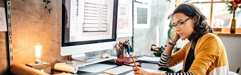 Eine junge Frau sitzt an einem Schreibtisch vor einem Monitor. Sie hält einen Stift in der Hand und blickt auf ein Blatt Papier.