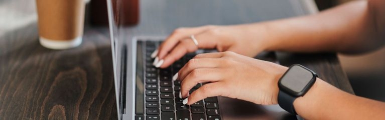 Hände tippen auf einer Tastatur, im Hintergrund steht ein Getränkebecher