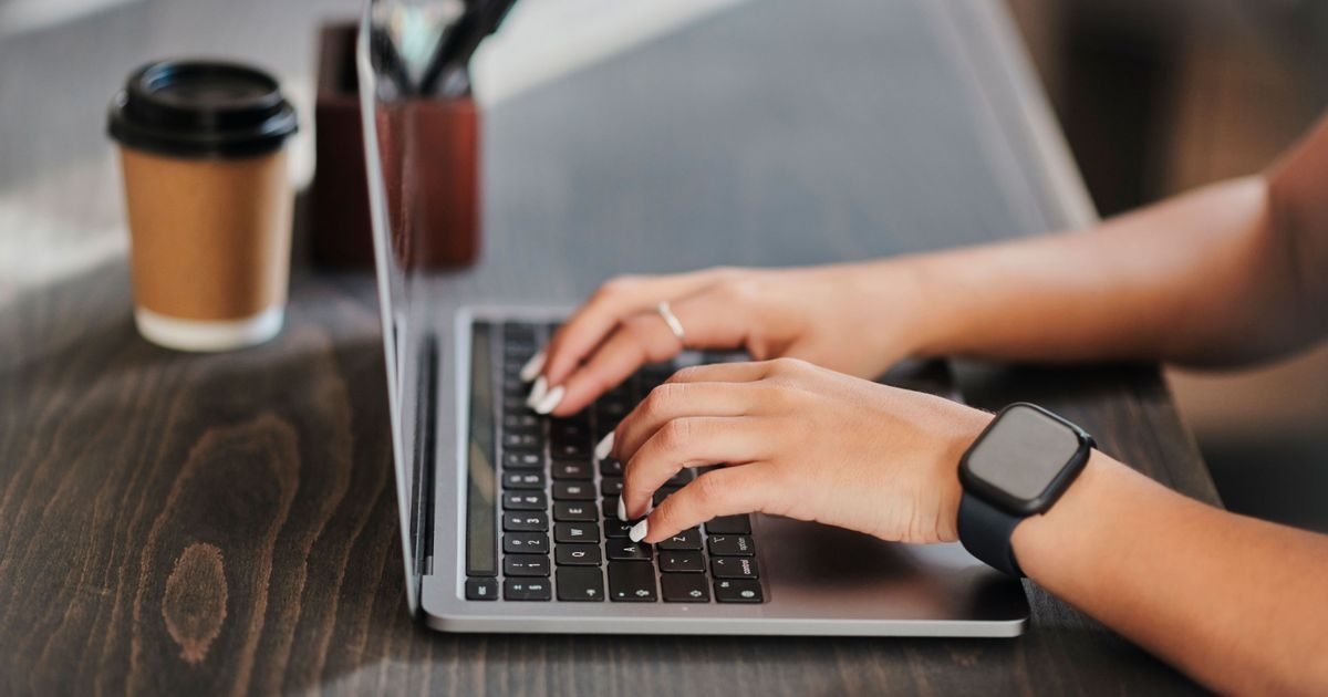 Hände tippen auf einer Tastatur, im Hintergrund steht ein Getränkebecher