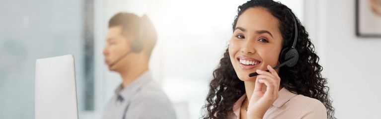 Eine Frau in einem Inbound-Call-Center sitzt an einem Schreibtisch vor einem Monitor. Sie telefoniert über ein Headset und lächelt dabei.