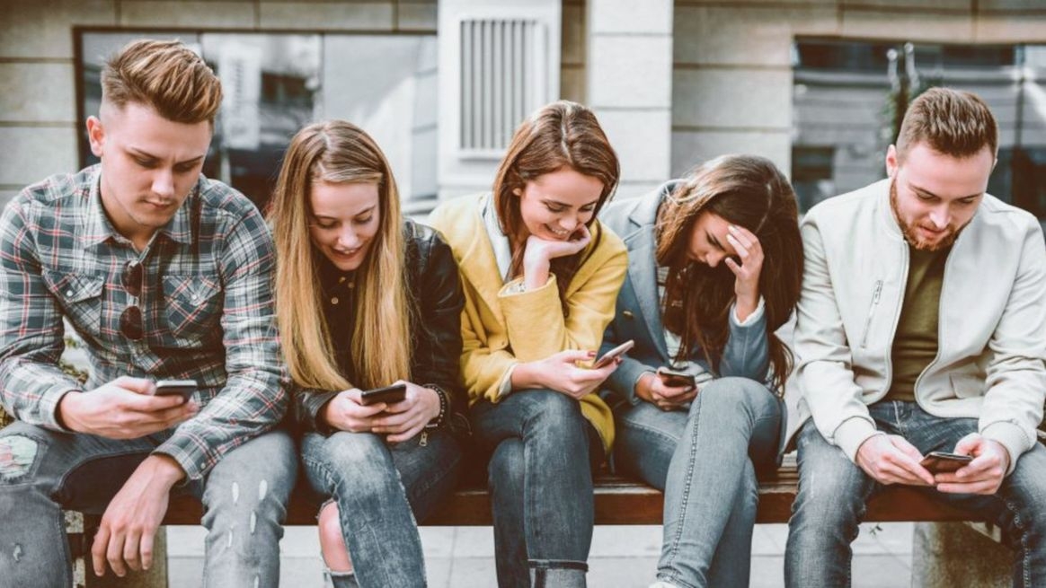Mehrere junge Menschen sitzen nebeneinander auf einer Bank und gucken auf ihre Smartphones