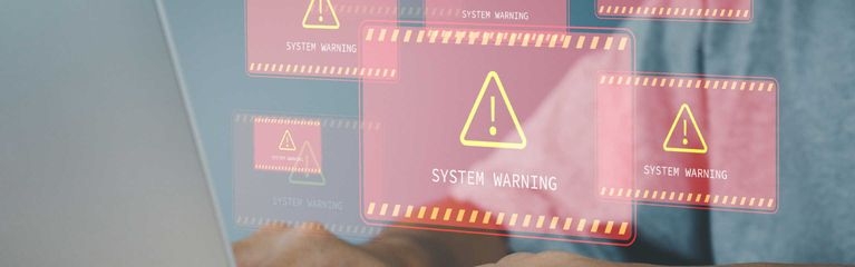 Zwei Hände auf der Tastatur eines Notebooks. Darüber rote digitale Hinweistafeln mit einem gelben Ausrufezeichen und dem Schriftzug System Warning