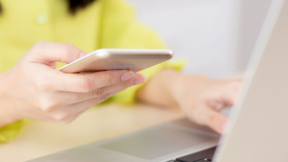 Eine Frau sitzt vor einem Laptop und hält in einer Hand ein Smartphone