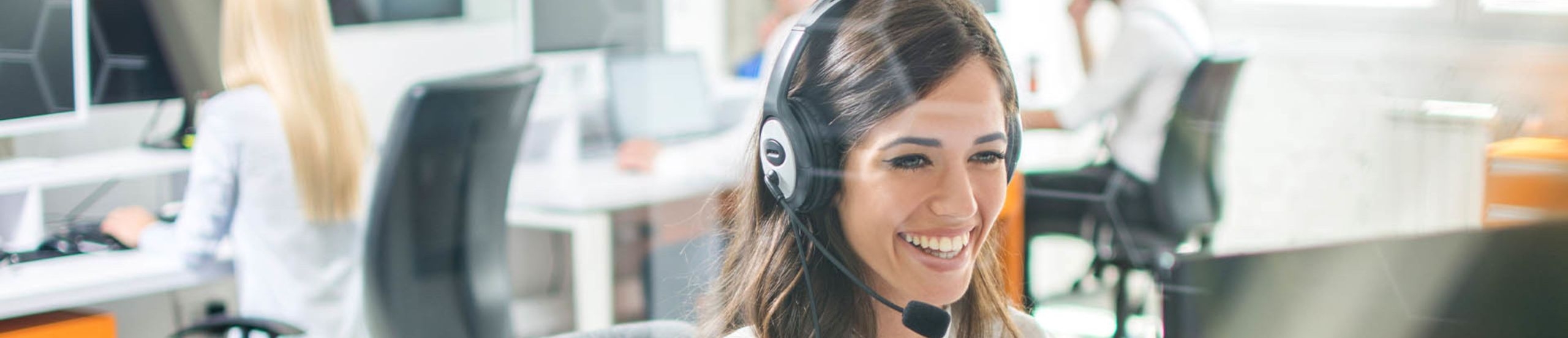 Lächelnde Frau sitzt im Büro an einem Computer und telefoniert via Headset