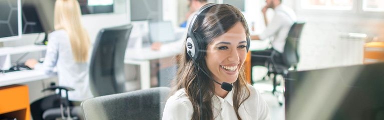 Lächelnde Frau sitzt im Büro an einem Computer und telefoniert via Headset