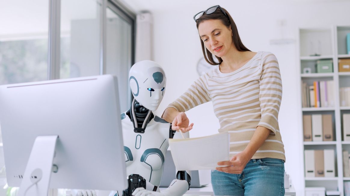 Eine Frau beugt sich am Arbeitsplatz über einen Roboter in Menschengestalt vor einem Computer. Sie zeigt diesem etwas auf Papier