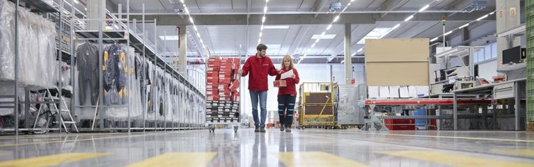 Ansicht einer Lagerhalle bei der Firma LOXXESS. 2 Mitarbeitende in roten Overalls besprechen ein Arbeitsblatt.