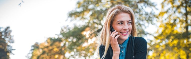 Eine Frau sitzt auf einer Parkbank und hält ein Handy am Ohr. Vor ihr auf der Parkbank ein aufgeklapptes Notebook.