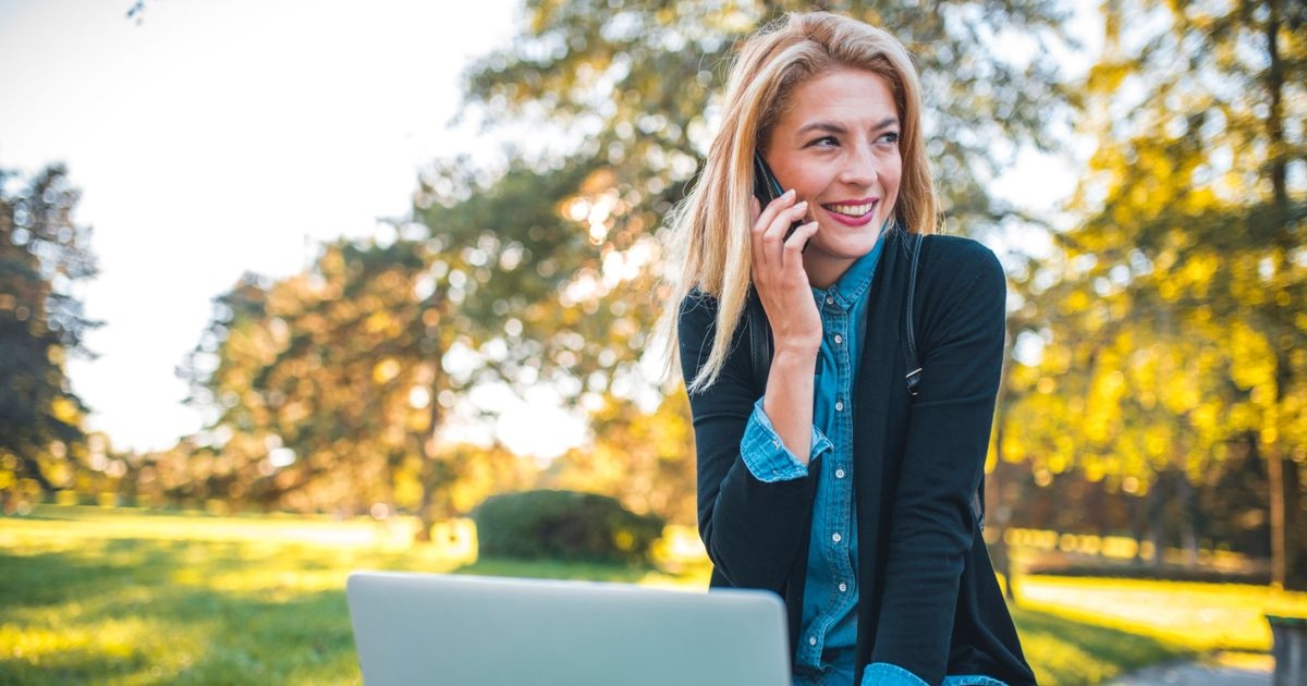 Eine Frau sitzt auf einer Parkbank und hält ein Handy am Ohr. Vor ihr auf der Parkbank ein aufgeklapptes Notebook.