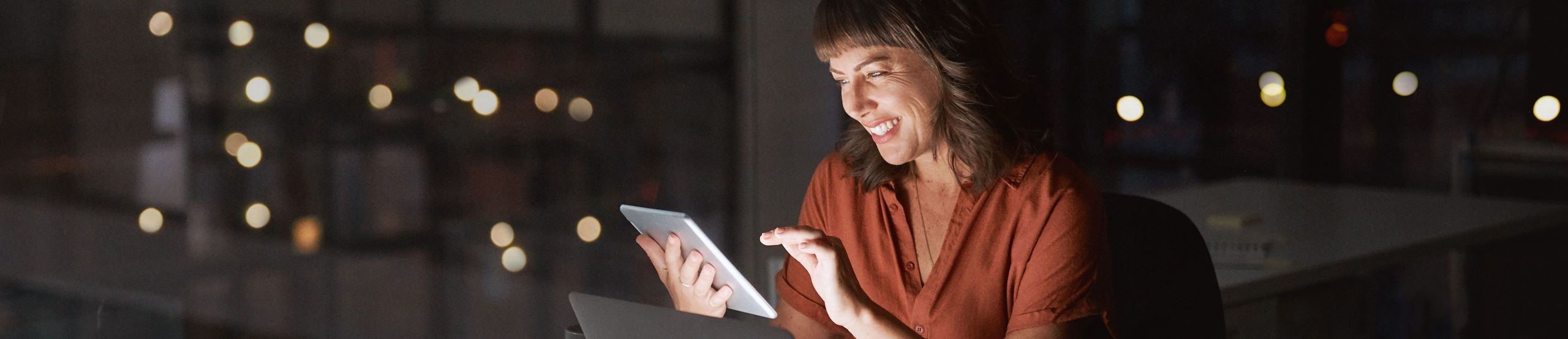 Frau sitzt im dunklen Büro und guckt lächelnd auf ein Tablet, das zusammen mit dem Notebook-Display die Szene beleuchtet