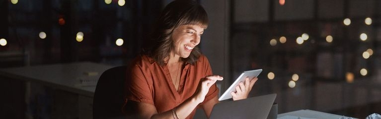 Frau sitzt im dunklen Büro und guckt lächelnd auf ein Tablet, das zusammen mit dem Notebook-Display die Szene beleuchtet