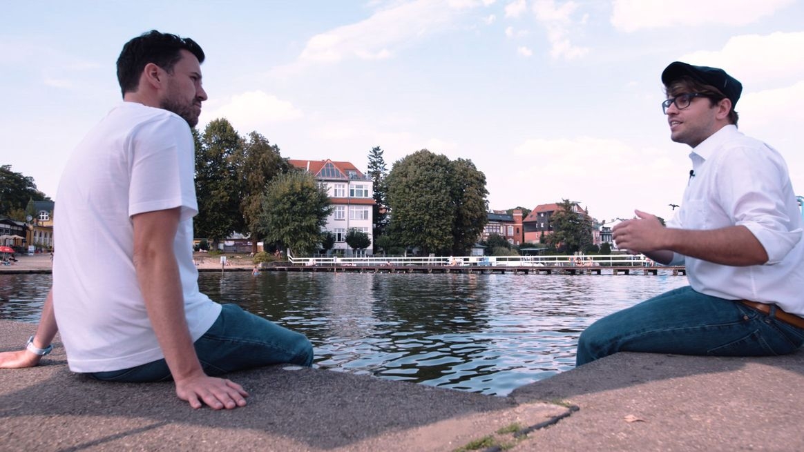 Max Wittrock und Karsten Kossatz sitzen am befestigten Ufer eines Gewässers. Im Hintergrund stehen Häuser auf der anderen Uferseite