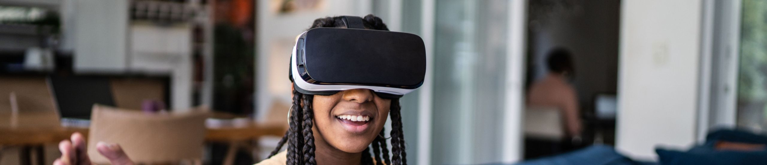 Eine junge Frau sitzt mit einem aufgesetzten VR-Headset in einem Zimmer und lächelt. 