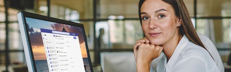 Eine junge Frau sitzt an einem Tisch vor einem Monitor. Darauf ist ein Chatfenster von Microsoft Teams zu sehen