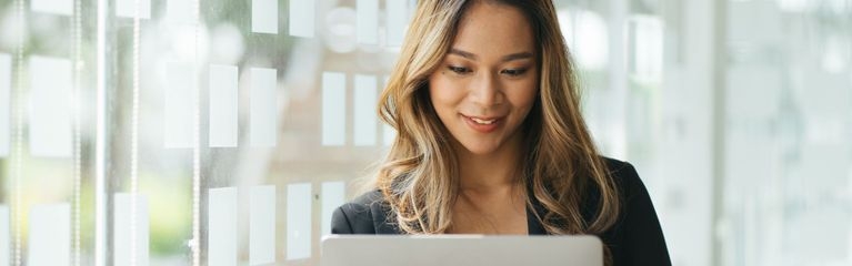 Frau steht in einem hellen Raum und guckt lächelnd auf ihr Notebook