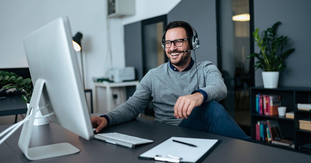 Lachender Mann mit Headset sitzt am Schreibtisch und guckt auf einen Bildschirm.