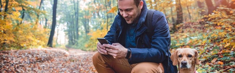 Mann hockt im herbstlichen Wald auf dem Boden und guckt auf ein Smartphone, neben ihm ein Hund