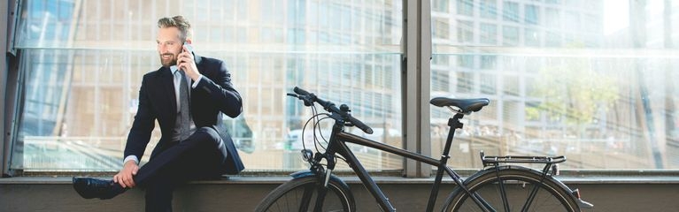 Mann im Anzug sitzt neben einem Fahrrad auf einem Sims und telefoniert