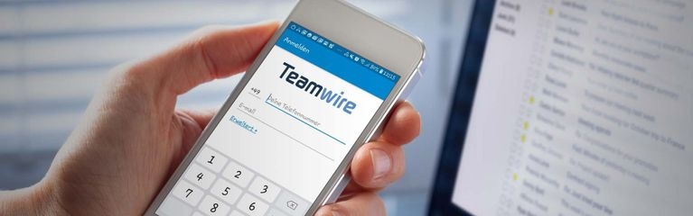 Eine Hand hält ein Smartphone über ein Notebook. Auf dem Display des Smartphones ist der Startbildschirm der Teamwire-App zu sehen