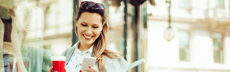 Frau mit Kaffeebecher in der rechten Hand schaut lächelnd auf ihr Smartphone in der linken Hand, vor einem Schaufenster stehend