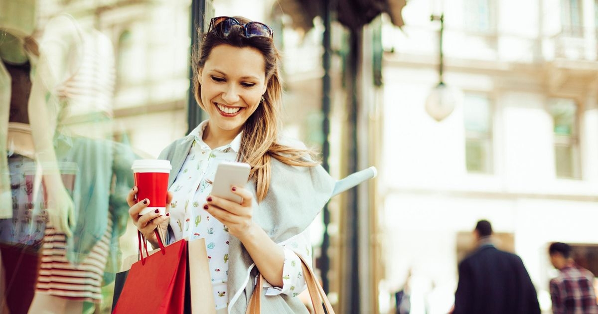 Frau mit Kaffeebecher in der rechten Hand schaut lächelnd auf ihr Smartphone in der linken Hand, vor einem Schaufenster stehend