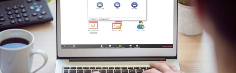Ein Notebook auf einem Tisch mit der Startseite von Zoom auf dem Bildschirm. Darauf liegen die Hände einer Person, daneben steht eine Tasse Kaffee auf dem Tisch.