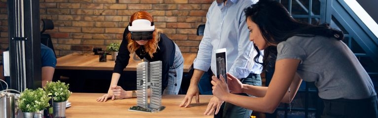 Drei Menschen arbeiten an einem Augmented Reality Projekt. Zwei von ihnen betrachten ein Architekturmodell durch eine AR-Brille, eine weitere Person fotografiert die Szene mit ihrem Tablet.