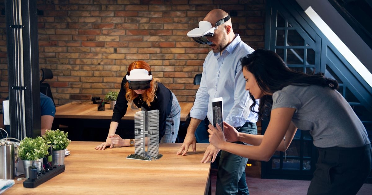 Drei Menschen arbeiten an einem Augmented Reality Projekt. Zwei von ihnen betrachten ein Architekturmodell durch eine AR-Brille, eine weitere Person fotografiert die Szene mit ihrem Tablet.