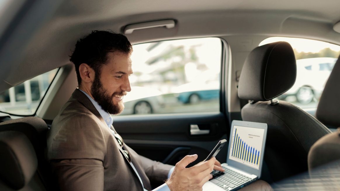 Mann auf Rückbank eines Autos guckt auf sein Handy in der rechten Hand, während die linke Hand auf der Laptop-Tastatur liegt
