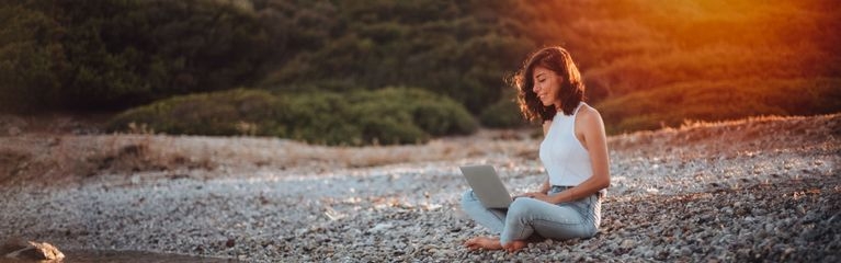 Eine junge Frau sitzt barfuß und lächelnd an einem Kiesstrand und hat ein Notebook auf dem Schoß Stock-Fotografie-ID:1257597625