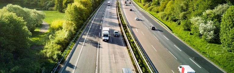 Eine dreispurige Straße mit Fahrzeugen von oben. Daneben sieht man Bäume und Büsche.