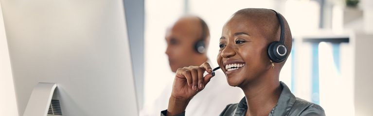 Eine Frau sitzt mit einem Headset vor einem Monitor und lächelt. Im Hintergrund sitzt ein Mann mit einem Headset.