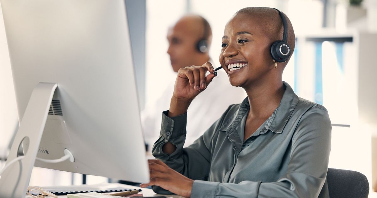 Eine Frau sitzt mit einem Headset vor einem Monitor und lächelt. Im Hintergrund sitzt ein Mann mit einem Headset.