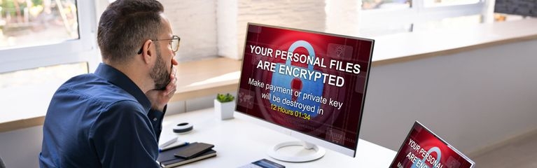 Ein Mann sitzt an einem Arbeitsplatz mit einem Bildschirm und einem Laptop. Auf beiden Bildschirmen ein Vorhängeschloss und der Text „Your personal files are encrypted“.