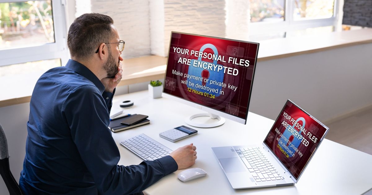 Ein Mann sitzt an einem Arbeitsplatz mit einem Bildschirm und einem Laptop. Auf beiden Bildschirmen ein Vorhängeschloss und der Text „Your personal files are encrypted“.