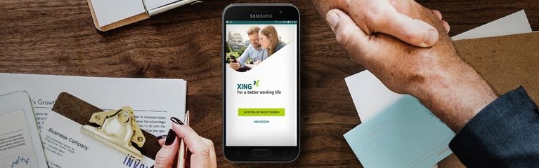 Die Hände von zwei Personen, die sich die Hand geben über einem Tisch mit Klemmbrettern, Dokumenten, einer Hand mit Stift und einem Smartphone mit dem Startbildschirm der Xing-App auf dem Display