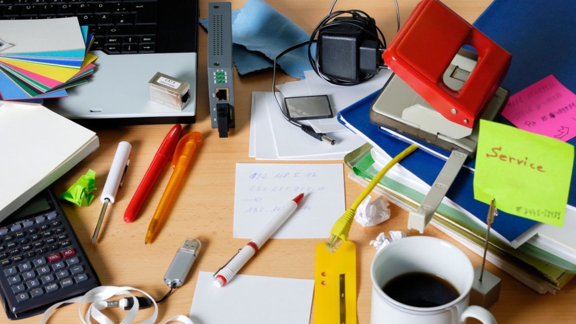 Unordentlicher Schreibtisch: Notebook, Stifte, Zettel, Kaffeetasse, Locher, Bücher und Müll liegen kreuz und quer übereinander
