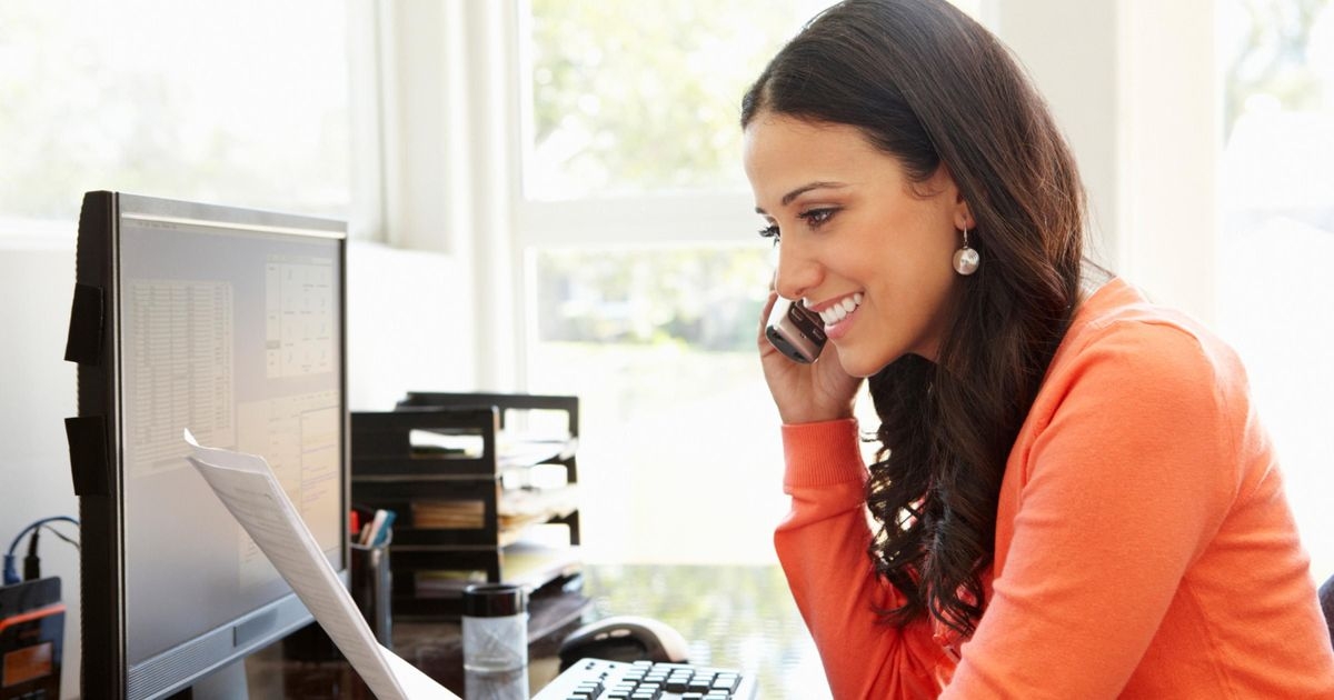 Eine Frau sitzt lächelnd an einem Schreibtisch vor einem Computer. In der rechten Hand hält sie ein Telefon ans Ohr, in der linken Papierblätter, die sie betrachtet.