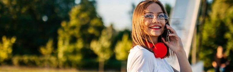 Eine junge Frau hält ein Smartphone ans Ohr und lächelt. Um den Hals trägt sie einen roten Kopfhörer, in der Hand hält sie einen Becher.