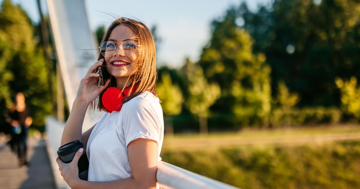 Eine junge Frau hält ein Smartphone ans Ohr und lächelt. Um den Hals trägt sie einen roten Kopfhörer, in der Hand hält sie einen Becher.