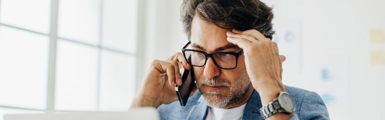 Ein Mann sitzt vor einem Notebook und hält Handy am rechten Ohr, linke Hand an linker Schläfe