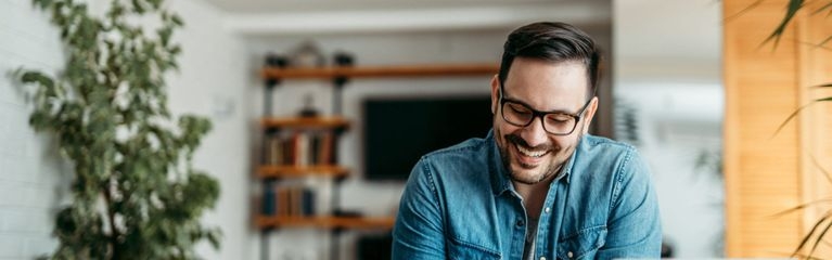 Ein Mann sitzt im Wohnzimmer vor dem Notebook und tippt lächelnd auf sein Smartphone