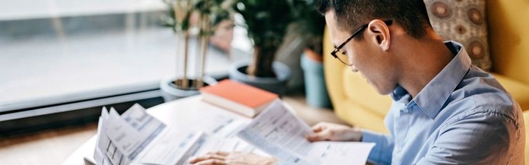 Ein Mann sitzt an einem Schreibtisch und betrachtet ein Dokument. Auf dem Tisch liegen weitere Dokumente und ein Taschenrechner.