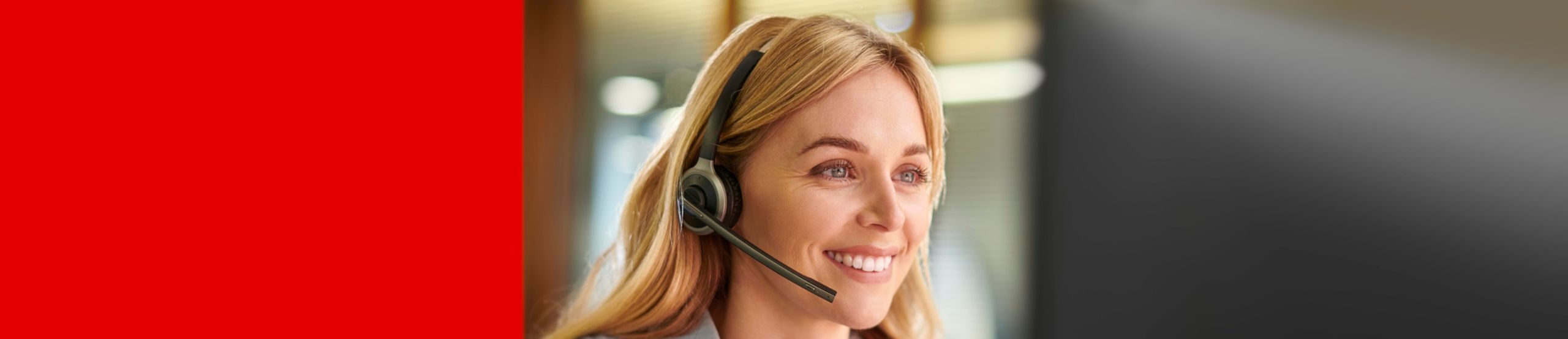 Eine Frau mit Headset sitzt lächelnd an einem Arbeitsplatz, im Hintergrund ein weiterer Mitarbeiter.