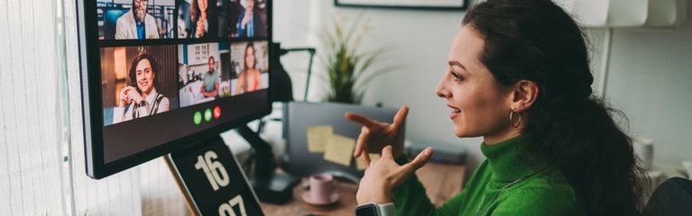 Eine junge Frau sitzt an einem Schreibtisch vor einem Bildschirm, auf dem ein virtuelles Meeting mit drei Frauen und drei Männern stattfindet.