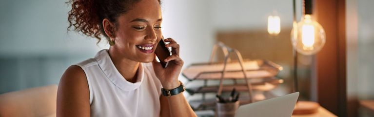 Eine junge Frau sitzt telefonierend an einem Schreibtisch und blickt auf ein Notebook